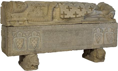  Fotografía de un sarcófago de piedra con la figura de un guerrero con su escudo reposando como losa superior, y los escudos familiares tallados en la base.